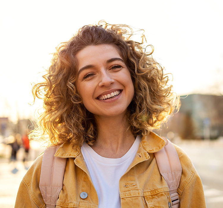 Smiling young woman with curly hair wearing a yellow jacket and backpack, outdoors in a bright setting, representing customer satisfaction and connection with GBIS Communications services.